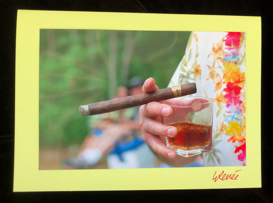 Close up of a man's hand holding a cigar and a glass of whiskey.  Man is wearing a Hawaiian shirt and a lei.